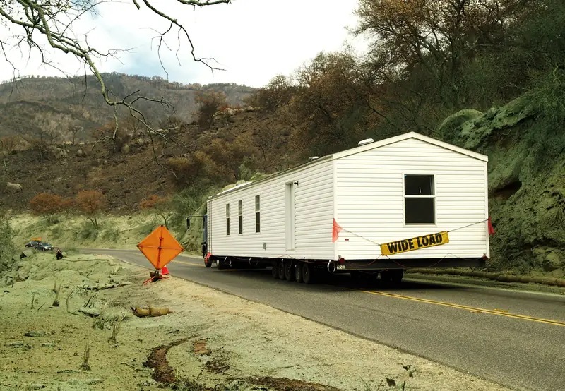A white single-section manufactured home on a tow rig with a WIDE LOAD banner, rolling along a two-lane road