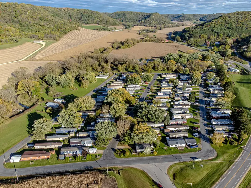 Aerial view of a manufactured home community with rows of homes along curving streets, surrounded by fields and wooded hills